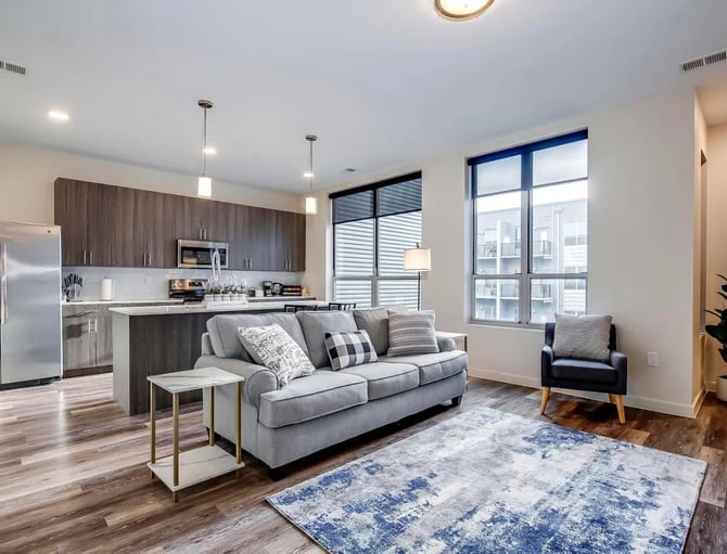 Open-plan living room and kitchen with gray sofa, dark wood cabinetry, stainless steel appliances, and large windows overlooking residential buildings.